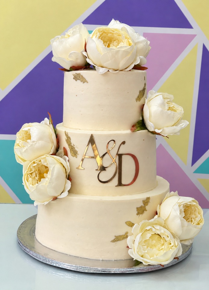Pastel de boda de tres pisos en color crema con flores blancas, detalles dorados e iniciales A y D